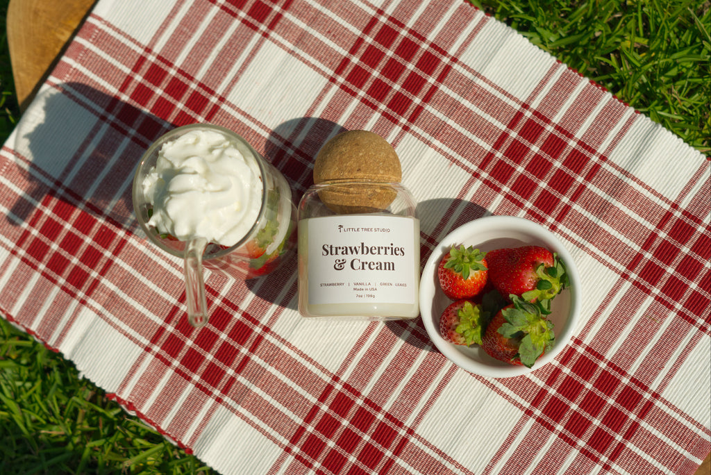 Strawberries and Cream candle with a glass of strawberry flavored desert and a bowl of strawberries on a red and white checkered tablecloth.