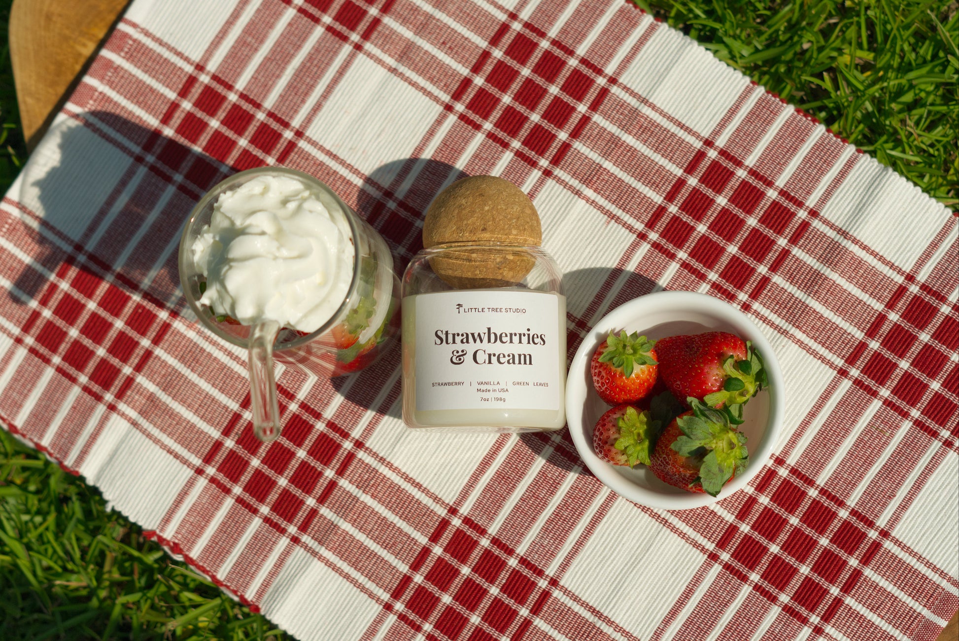Strawberries and Cream candle with a glass of strawberry flavored desert and a bowl of strawberries on a red and white checkered tablecloth.